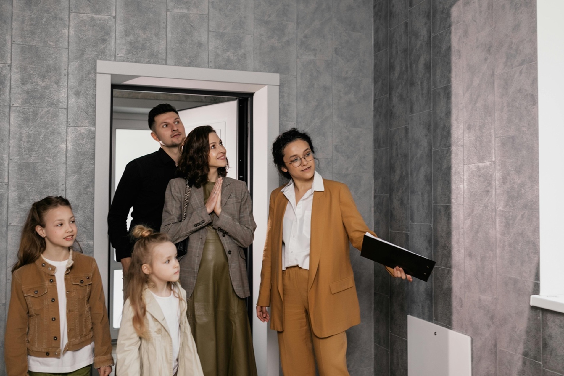 a woman in a burnt orange suit showing a family of four through a house with grey walls